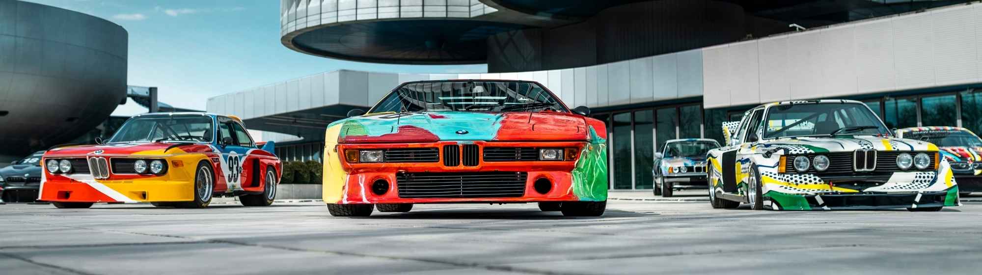 Trois BMW Art Cars exposées devant des bâtiments modernes, mettant en valeur des voitures de course colorées.
