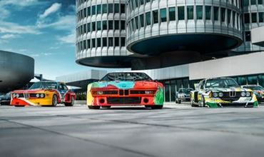 Trois BMW Art Cars exposées devant des bâtiments modernes, mettant en valeur des voitures de course colorées.