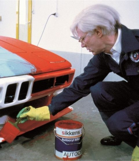 Andy Warhol peignant une voiture BMW dans un atelier, avec une touche de peinture rouge vif.